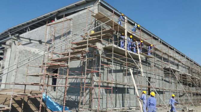 This image shows construction workers in blue overallas and yellow hard hats working on an incomplete cement/concrete structure that is covered by scaffolding